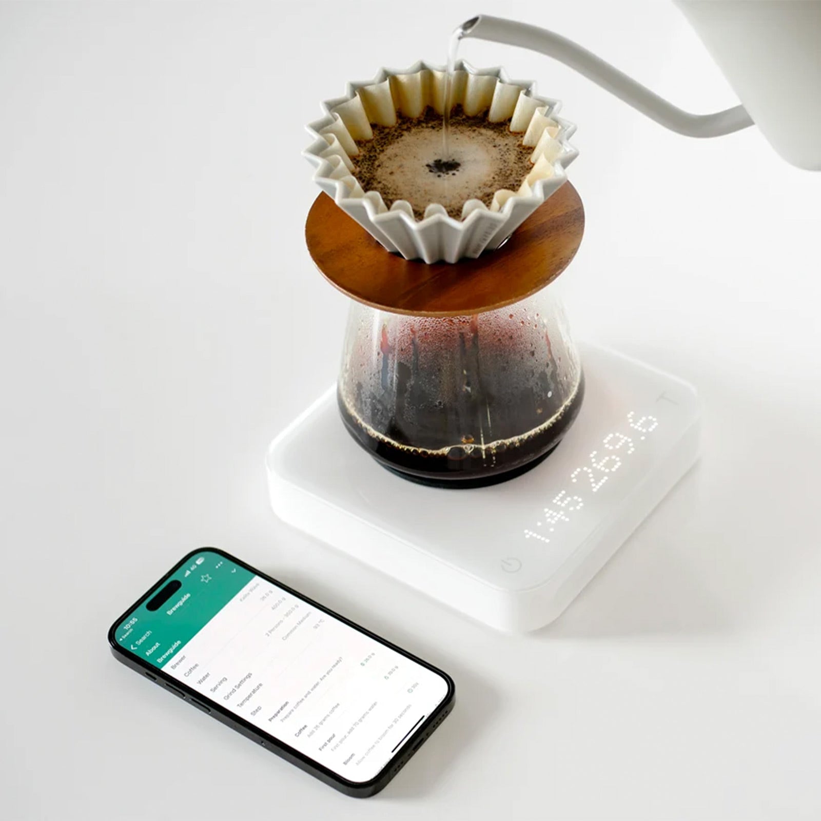 A pour-over coffee setup on a Acaia Pearl S Scales White digital scale, with hot water being poured from a white gooseneck kettle into a white paper filter cone over dark coffee in a glass carafe.