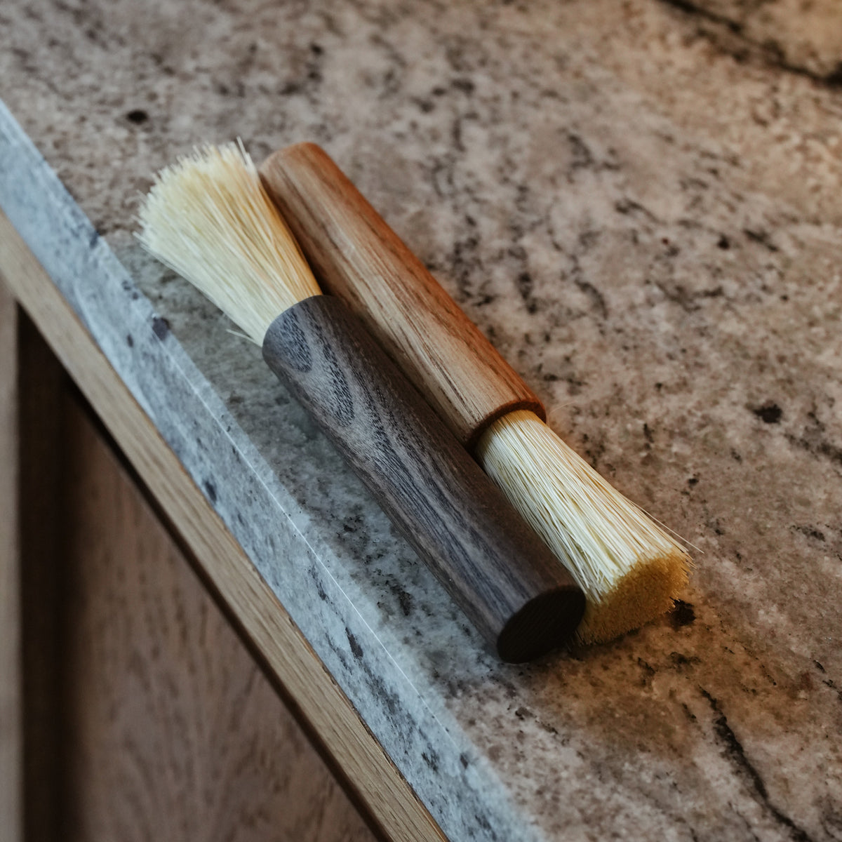 Two small coffee brushes with natural bristles, one featuring a dark wood handle and the other a light wood handle, shown on a kitchen counter.