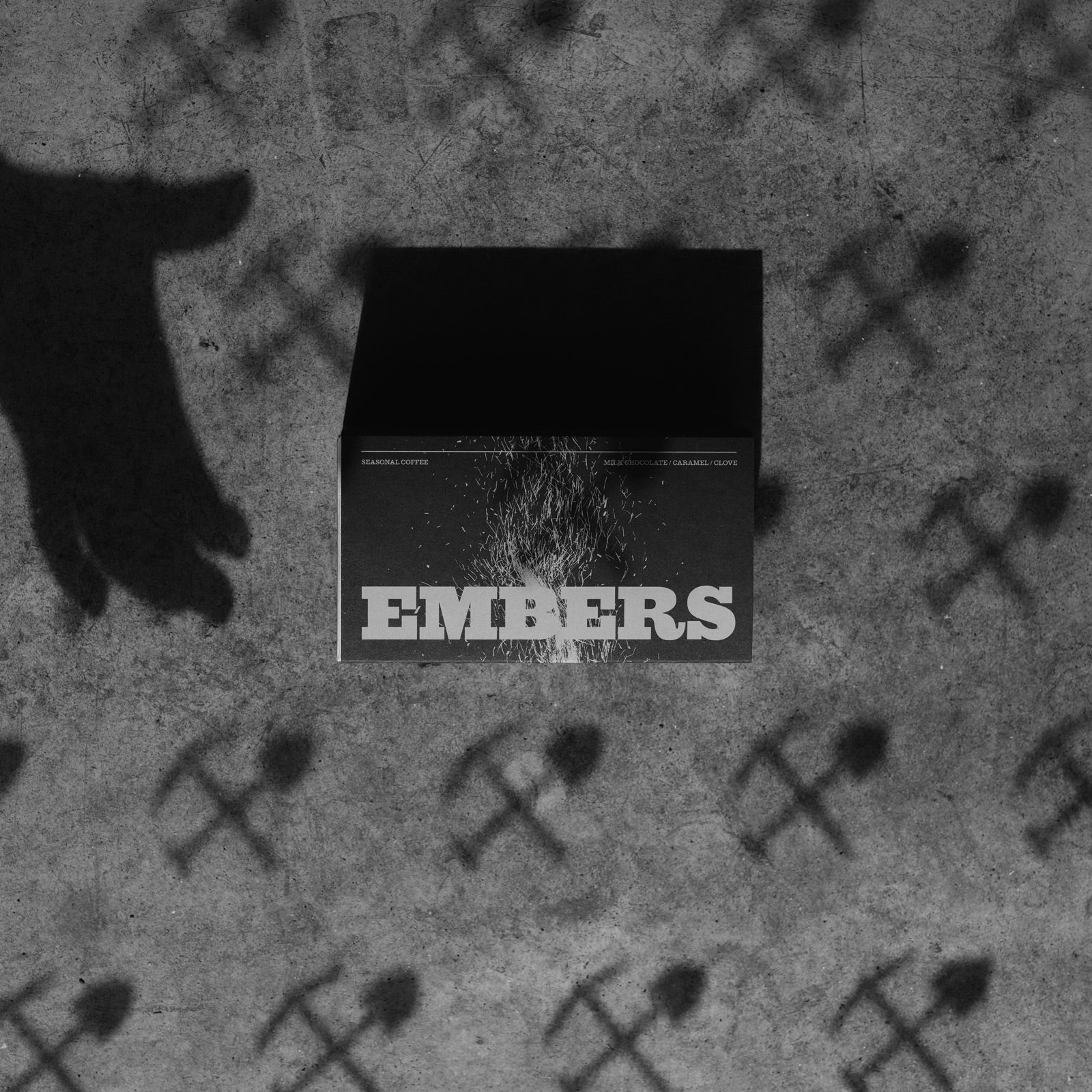 A black rectangular box or package with the word 'EMBERS' in large white letters, placed on a textured gray surface. The scene is monochrome, with a large shadow cast across the left side and repeated hammer-and-sickle symbols printed across the background.