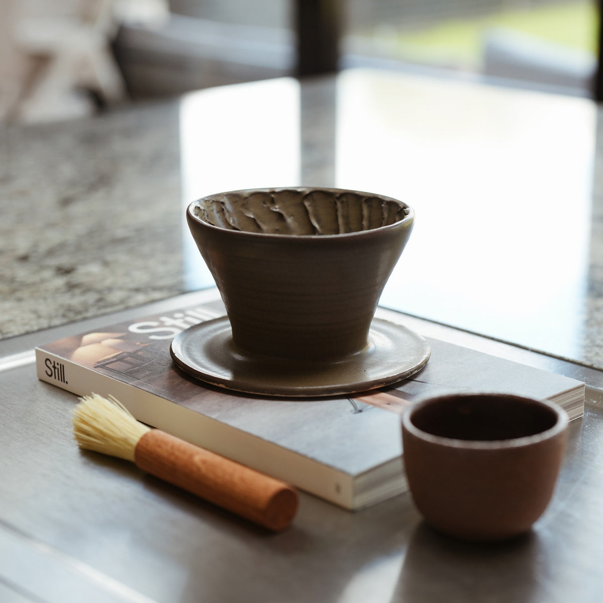 Ash black V60 ceramic hand made pottery coffee with a oak coffee brush and brown ceramic espresso cup on a kitchen work top
