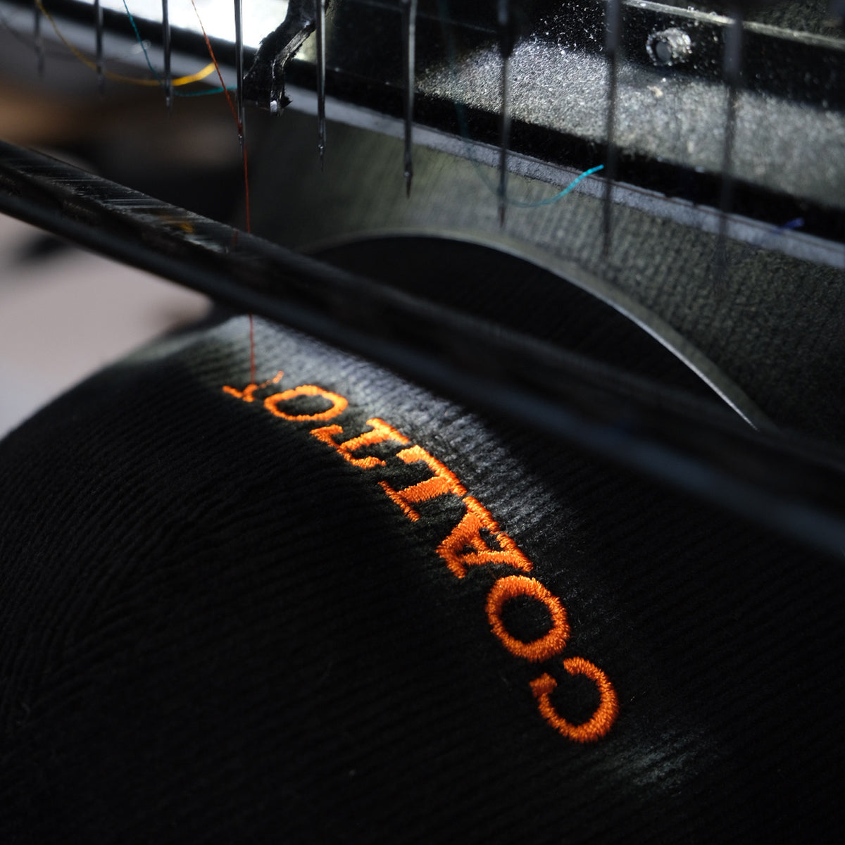 Close-up of a black textured fabric item with orange embroidered lettering reading 'COALITION' under a needle or embroidery thread, with dark machinery visible in the background.