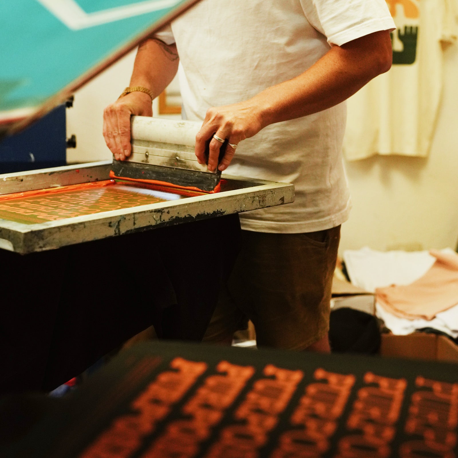 A person wearing a white t-shirt and brown shorts is using a screen printing press to apply orange ink onto a white fabric. The scene is shot from an overhead angle in a warm, yellow-toned indoor workshop environment.