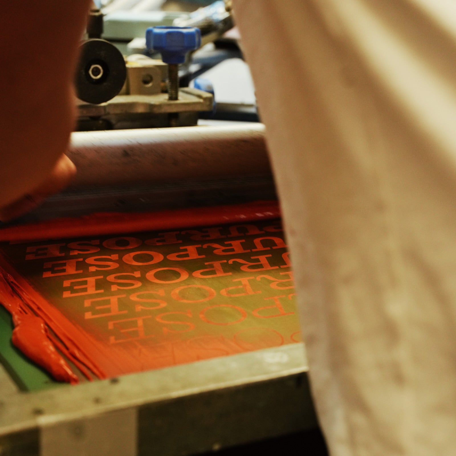 A close-up, angled view of a screen printing press in action, with a person's hand guiding a roller over orange-red ink on a sheet of text that reads 'PURPOSE' upside down.