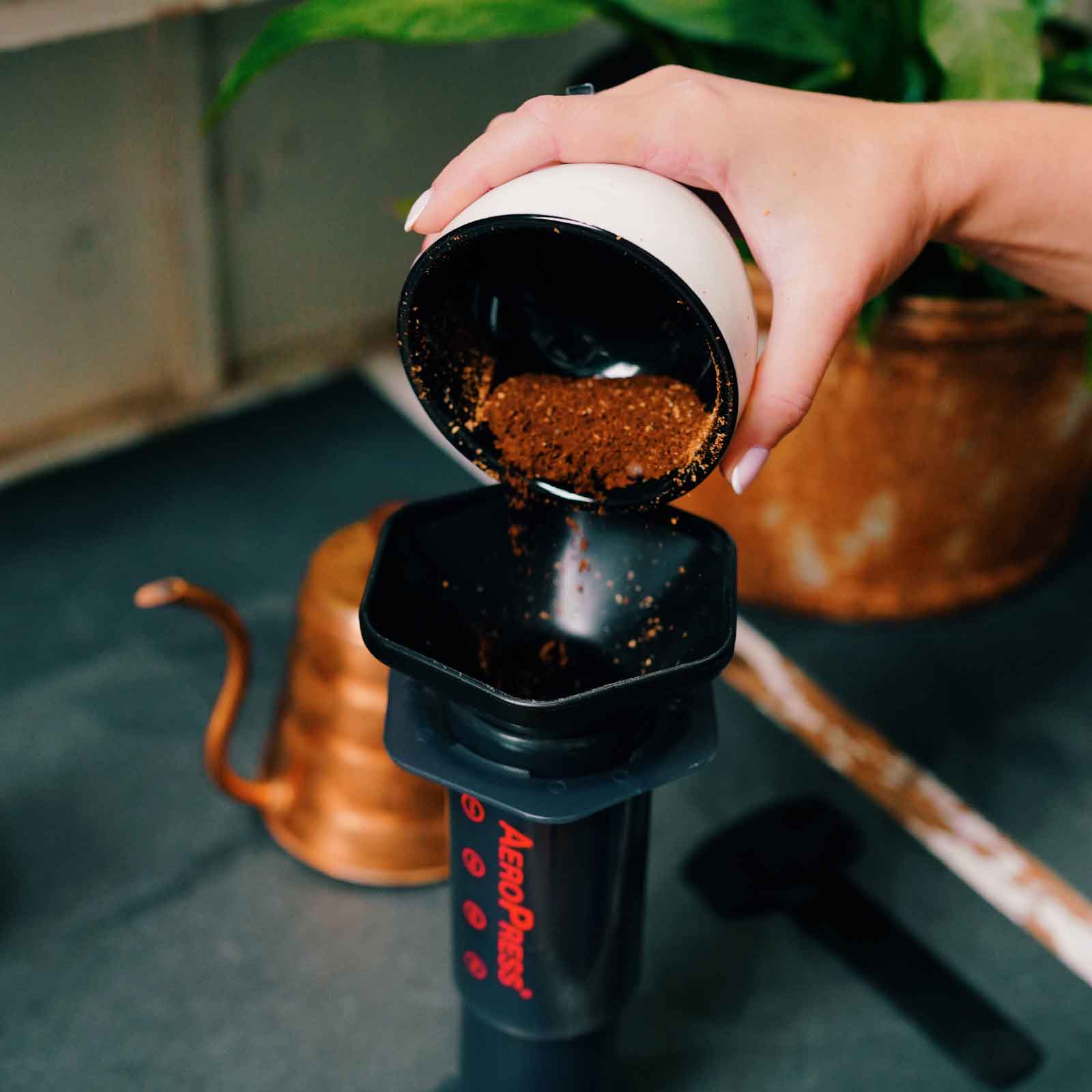 Hand pouring coffee grounds into a black AeroPress coffee maker with a blurred background.