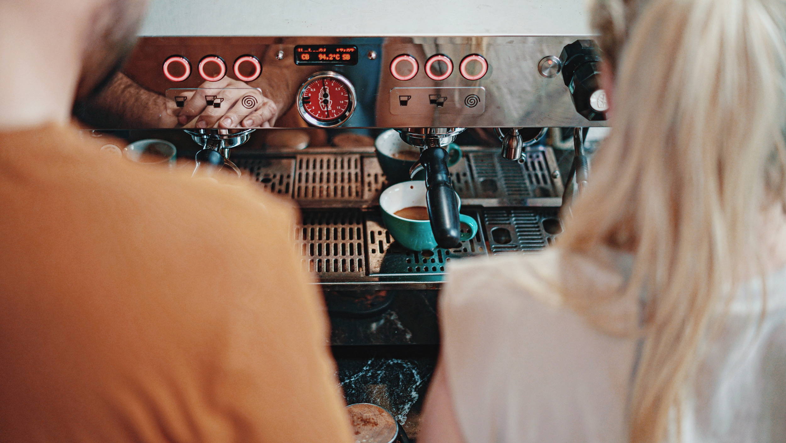 Two people looking at a la marzocco coffee machine waiting for the espresso pouring into a blue mug to finish