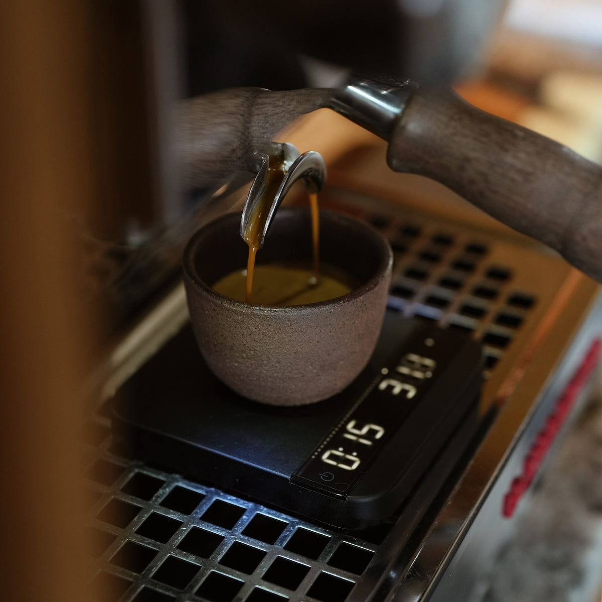 A close-up of espresso being poured from a metal portafilter into a small speckled brown ceramic cup placed on a digital scale on a la marzocco coffee machine