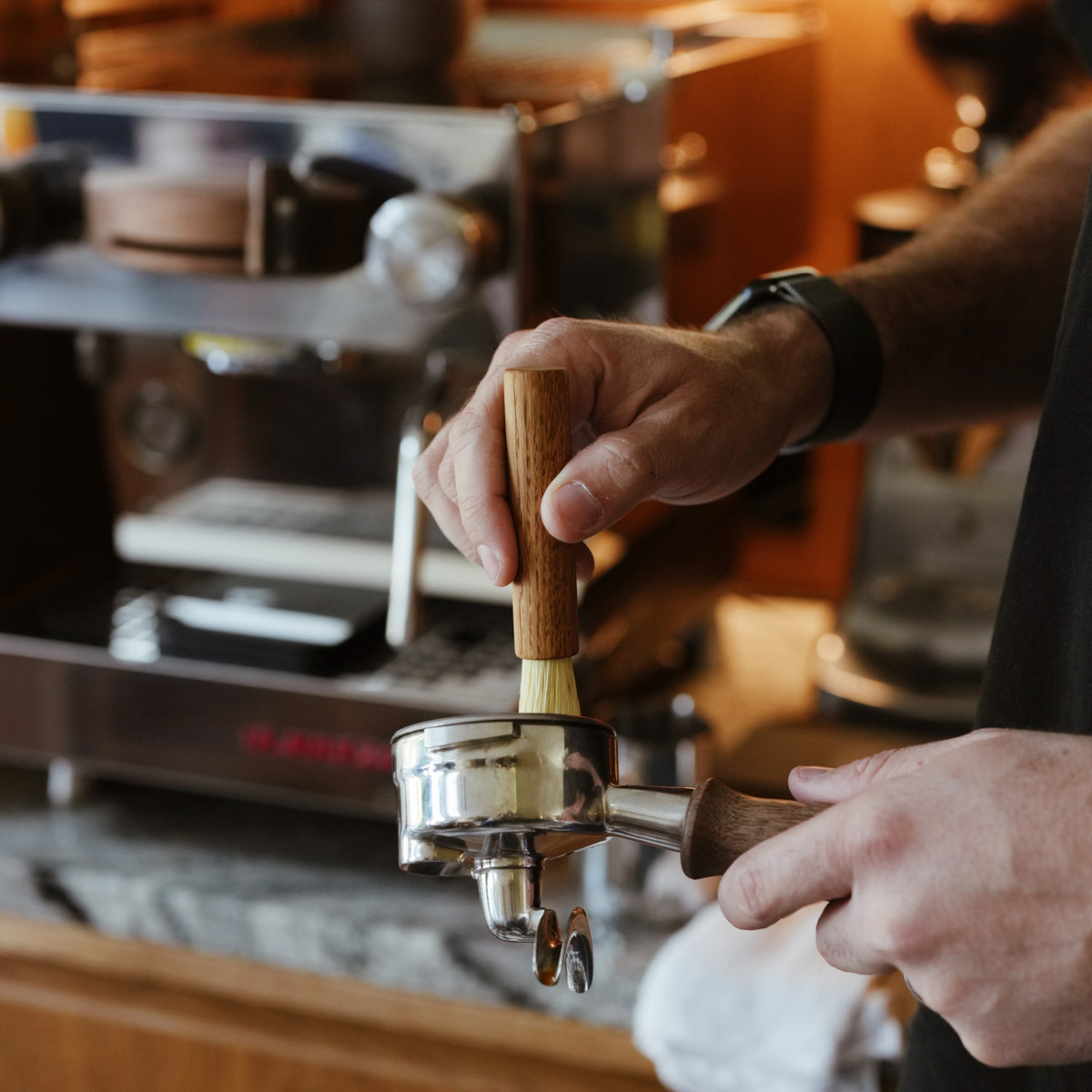 Small Oak coffee brush with natural bristles and a wooden handle. Shown in a kitchen, person cleaning out a La Marzocco coffee machine portafilter.
