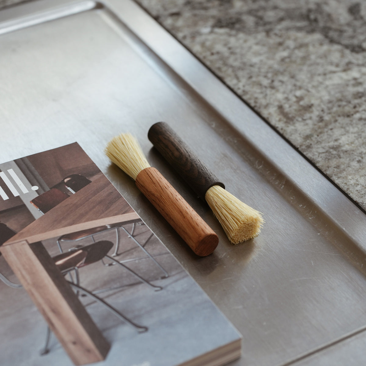 Two small coffee brushes with natural bristles, one featuring a dark wood handle and the other a light wood handle, shown on a plain stainless steel kitchen counter.