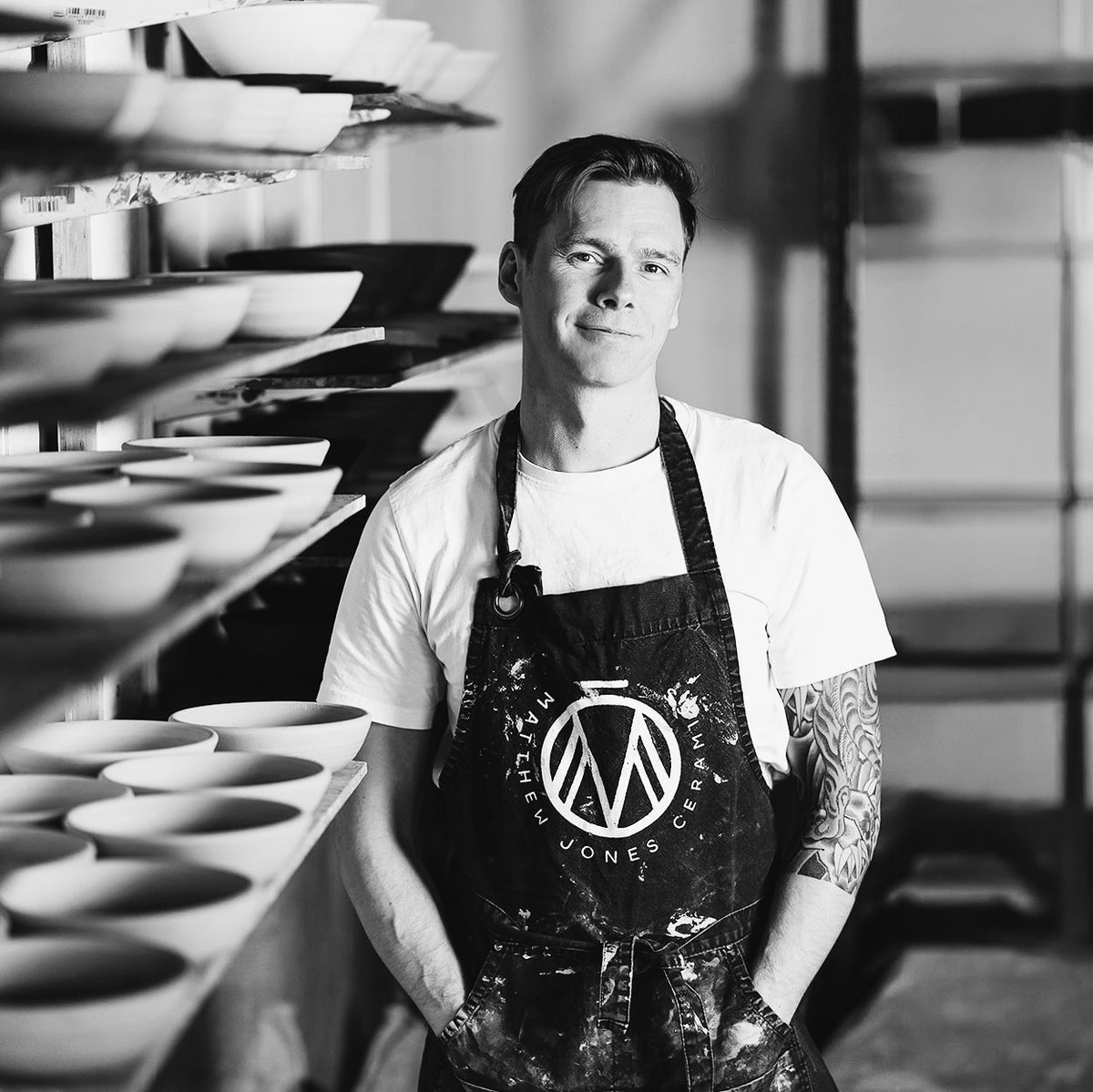 A black and white portrait of a man wearing a white t-shirt and a dark apron with a circular 'Marble Jones Ceramics' logo, standing in a pottery studio with shelves of ceramic bowls in the background.