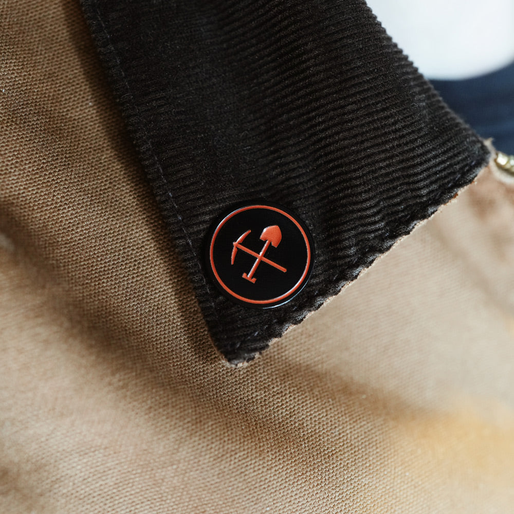 Close-up of a black corduroy lapel collar with a circular black enamel pin featuring a red-orange pickaxe and shovel symbol, set against a beige fabric background.