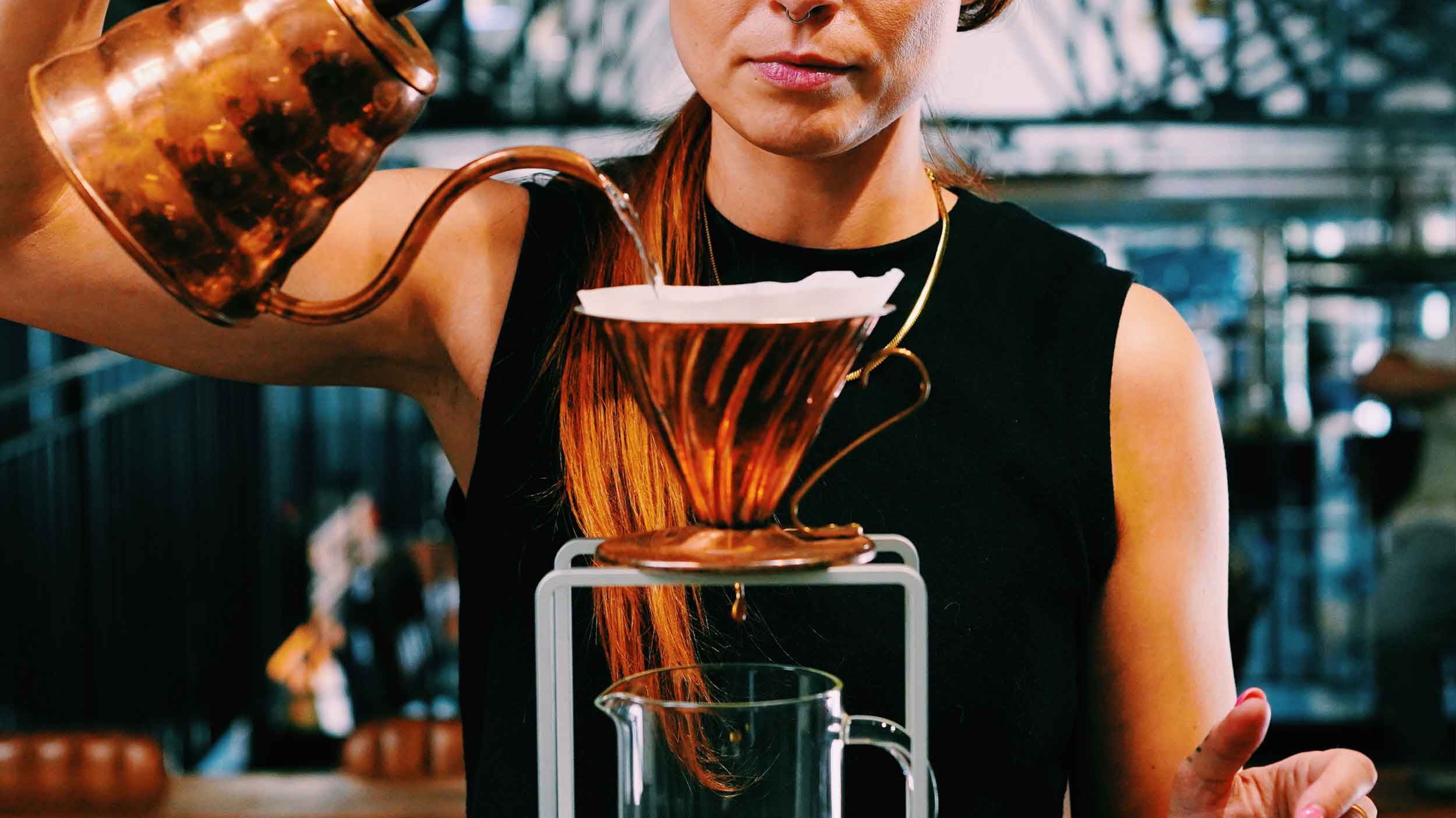 Person pouring coffee from a bronze kettle and V60 into a glass carafe.