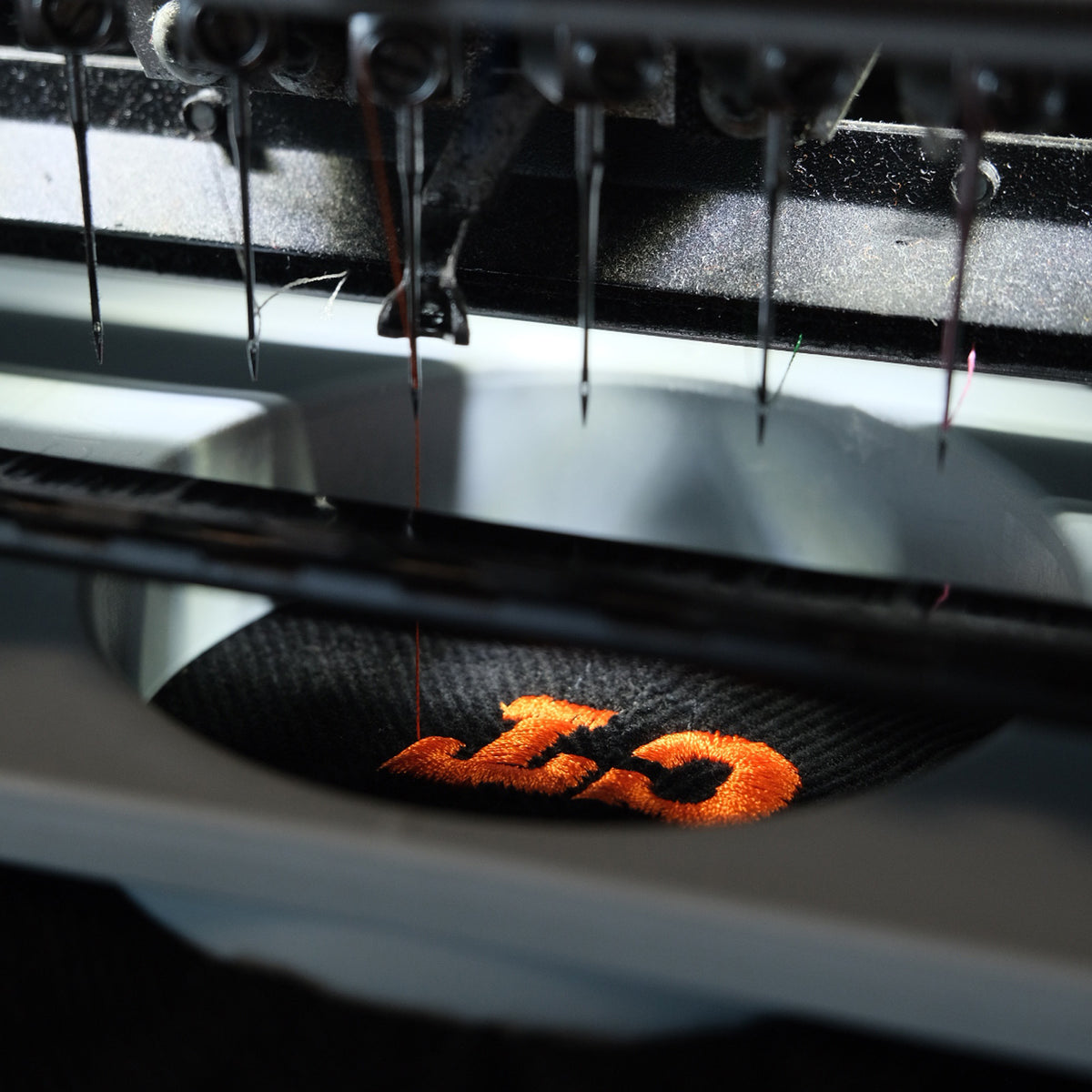 Close-up of an embroidery machine needle stitching an orange embroidered logo onto a black fabric surface.